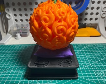 May include: An orange, flame-like fruit with a green stem and curly tendrils, resting on a purple pillow and a black base. The fruit is the focal point, showcasing intricate details. The base has two circular designs. The background is a work surface.