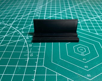 May include: A small, black, rectangular object with a raised back and a grooved base. The object is made of a solid material and sits on a green cutting mat with white grid lines and measurements in inches.