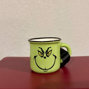 May include: A green ceramic mug with a black rim and handle. The mug features a black and yellow illustration of the Grinch's face.