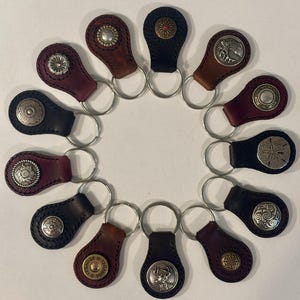 Puede incluir: Una colección de llaveros de cuero en varios colores, incluyendo negro, marrón y burdeos. Cada llavero presenta un botón metálico decorativo con diseños intrincados. Los llaveros están dispuestos en un patrón circular.