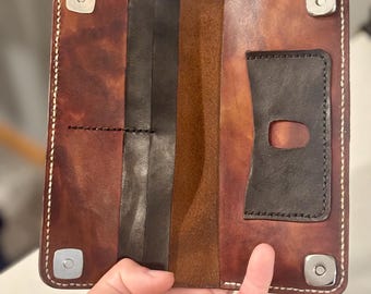 May include: Open, handmade leather wallet in shades of brown and black. The wallet features multiple compartments, including a card slot and a billfold. It is secured with four silver snap closures and white stitching.
