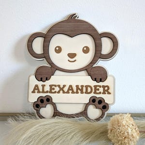 Könnte beinhalten: Holzschild in Affenform mit dem Namen "ALEXANDER" in Großbuchstaben. Der Affe ist braun und beige, mit einem lächelnden Gesicht und hält ein rechteckiges Schild. Das Schild ist ein dekorativer Gegenstand.