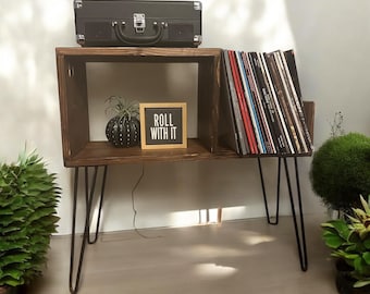 May include: A dark brown wooden record player stand with black hairpin legs. The stand has a compartment for records and a top shelf with a black record player case. A framed sign reads "ROLL WITH IT".