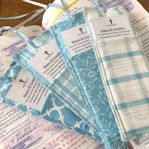 May include: A collection of handmade fabric bookmarks in various light blue patterns, each with a coordinating ribbon. The bookmarks feature a printed label with the text "Peace & Comfort" and a religious cross symbol. The bookmarks are displayed on top of an open book.