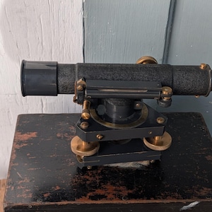 May include: A vintage surveying level instrument featuring a black textured cylindrical telescope, a central spirit level vial, and a dark gray base with three adjustable brass leveling screws and a circular rotating dial for precise alignment.