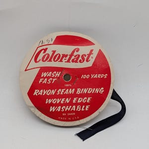 May include: A circular spool of black rayon seam binding with a red and white paper label. The label reads Colorfast, 100 Yards, Rayon Seam Binding, Woven Edge, Washable, 100%, RN 24429, and Made in USA. A single length of black ribbon extends from the spool.