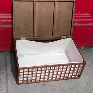 May include: A wooden storage chest with a hinged lid, open to reveal a white fabric interior. The chest has a lattice-style front and sides, and is set against a red background.