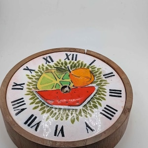May include: A round wall clock with a wooden frame and a white ceramic face. The face features black Roman numerals and a central illustration of a sliced watermelon, a lime wedge, and an orange, all surrounded by a green leaf pattern.