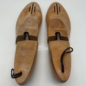 May include: Pair of wooden shoe trees. The shoe trees are light brown with a dark brown band and a dark brown pull cord. The shoe trees have a classic design with ventilation slits on the top.