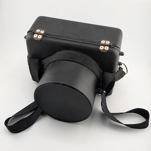 Può includere: Custodia per fotocamera nera con attacco per obiettivo cilindrico. La custodia è rettangolare con angoli arrotondati e cerniere color ottone. È attaccata una cinghia nera per il trasporto. La custodia è realizzata in materiale resistente.