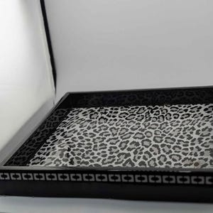 May include: A rectangular black tray with a glossy finish. The interior features a leopard print pattern in shades of gray and white. The tray's border is decorated with a repeating geometric design.