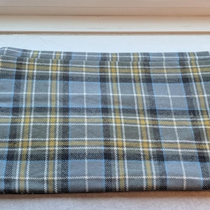 May include: A folded rectangular fabric with a plaid pattern. The pattern features a gray base with intersecting lines in light blue, yellow, white, and black. The fabric is likely used for sewing or crafting projects.