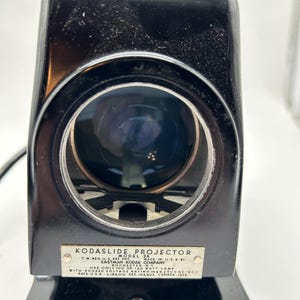 May include: A vintage Kodaslide Projector, Model 2A, in black. The projector has a large lens and a metal plate with text that reads "Kodaslide Projector" and other details. The device is made by Eastman Kodak Company.