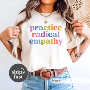 May include: Ivory-colored t-shirt with the words "practice radical empathy" in rainbow-colored letters. The shirt has a crew neck and short sleeves. The model is wearing a brown belt with a heart-shaped buckle and blue jeans.