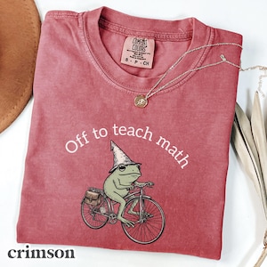 May include: Crimson-colored t-shirt with the phrase "Off to teach math" and a graphic of a frog riding a bicycle. The frog wears a wizard hat and carries a satchel. A gold necklace with a pendant rests on the shirt.