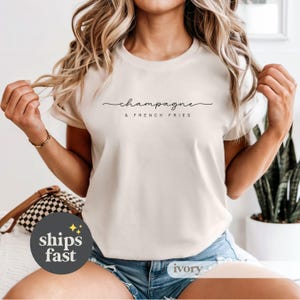 May include: Ivory-colored t-shirt with the words "champagne & french fries" in a black script font. The shirt has short sleeves and a crew neck. The model is wearing blue denim shorts.