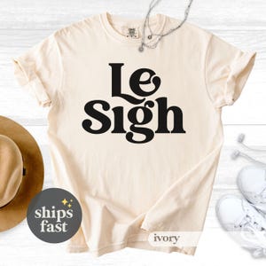 May include: Ivory-colored t-shirt with the words "Le Sigh" in bold, black letters. The shirt has a crew neck and short sleeves. A silver necklace and a hat are visible in the background. The shirt is labeled "ivory".