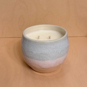 May include: A two-wick candle in a ceramic container. The container has a light blue and pink ombre effect. The candle wax is white, and the wicks are visible. The candle sits on a light brown surface.