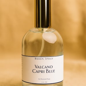 Könnte beinhalten: Eine transparente Raumduftflasche mit goldenem Verschluss. Das Etikett trägt die Aufschrift "Room Spray" und "Valcano Capri Blue" mit dem Text "By Hudson Park". Die Flasche enthält 148 ml Produkt.