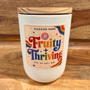 May include: A white candle vessel with a wooden lid featuring a label that reads HUDSON PARK Fruity + Thriving with a disco ball, rainbow accents, and stars. The label also lists the scents lemon, mint, vanilla, and berry in black text.