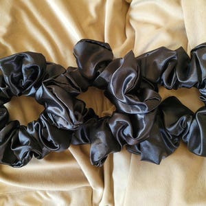 May include: Three black satin scrunchies, a popular hair accessory, are arranged on a beige fabric background.