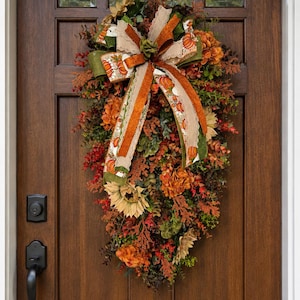 May include: A teardrop-shaped autumn wreath featuring artificial sunflowers, orange and brown foliage, and green leaves. A large decorative bow with a pumpkin print, burlap texture, and green edges is centered at the top of the floral arrangement.