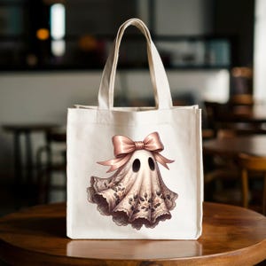 May include: A white canvas tote bag featuring a whimsical ghost design. The ghost is adorned with a pink bow and a delicate lace-like fabric with floral accents. The bag has sturdy handles and is set against a blurred background.
