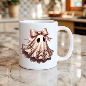 May include: White ceramic mug featuring a whimsical ghost design. The ghost is adorned with a pink bow and a draped, floral-patterned veil. The mug has a curved handle and is suitable for hot or cold beverages.