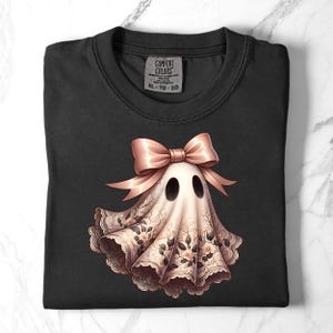 May include: Black t-shirt featuring a ghost design. The ghost is adorned with a pink bow and a floral lace-like pattern. The shirt has a crew neck and a tag that reads "COMFORT COLORS XL-TG-EG".