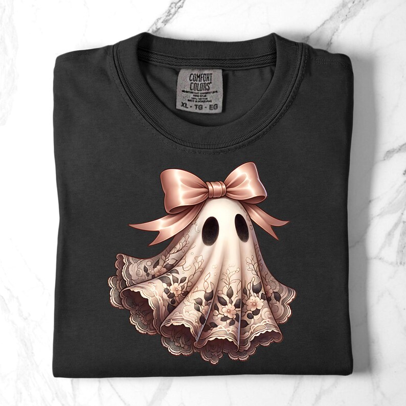 May include: Black t-shirt featuring a ghost design. The ghost is adorned with a pink bow and a floral lace-like pattern. The shirt has a crew neck and a tag that reads "COMFORT COLORS XL-TG-EG".
