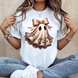 May include: White t-shirt featuring a whimsical ghost design. The ghost is adorned with a pink bow and a floral lace-like fabric. The design has two black eye holes. A unique and stylish Halloween-themed apparel.