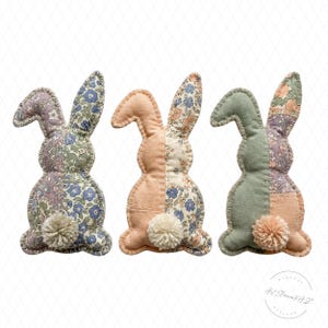 May include: Three handmade fabric bunny decorations. Each bunny has a pom-pom tail and is made of patchwork fabric in pastel colors, including pink, lavender, and green. The ears are upright, and the bunnies are stitched together.