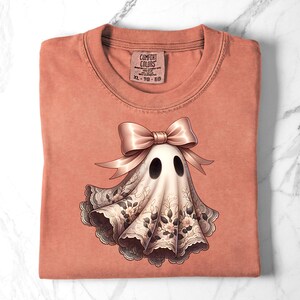 May include: A coral-colored t-shirt with a graphic of a ghost wearing a bow and lace. The ghost has two black eyes and a white body with floral lace trim. The tag reads "COMFORT COLORS" and "XL - TG - EG".