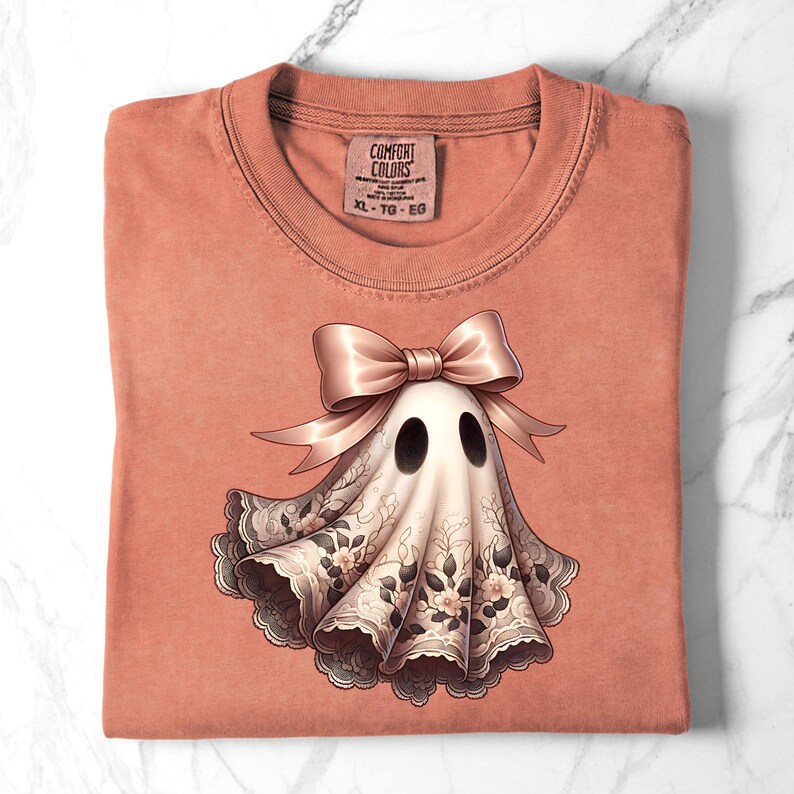 May include: A coral-colored t-shirt with a graphic of a ghost wearing a bow and lace. The ghost has two black eyes and a white body with floral lace trim. The tag reads "COMFORT COLORS" and "XL - TG - EG".