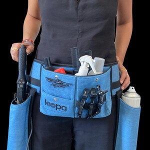 May include: A blue denim styling belt with multiple pockets and loops for holding hair styling tools. The belt has a black strap and a black buckle. The belt is worn by a person and has the text "loopa hair no assistant  just skills" printed on it.
