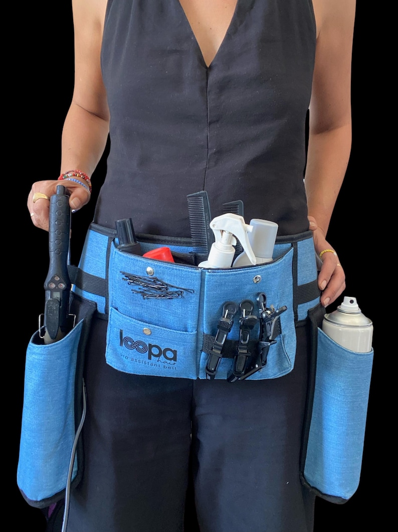 May include: A blue denim styling belt with multiple pockets and loops for holding hair styling tools. The belt has a black strap and a black buckle. The belt is worn by a person and has the text "loopa hair no assistant  just skills" printed on it.