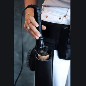 May include: A black leather styling tool holder with a black curling iron inside. The holder is attached to a black belt worn around the waist.