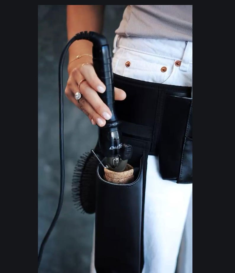 May include: A black leather styling tool holder with a black curling iron inside. The holder is attached to a black belt worn around the waist.