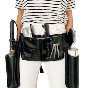 May include: Black leather stylist belt with multiple pockets and holders for tools, including a curling iron, shears, combs, and hairspray. The belt is worn by a person with white pants and a striped shirt.