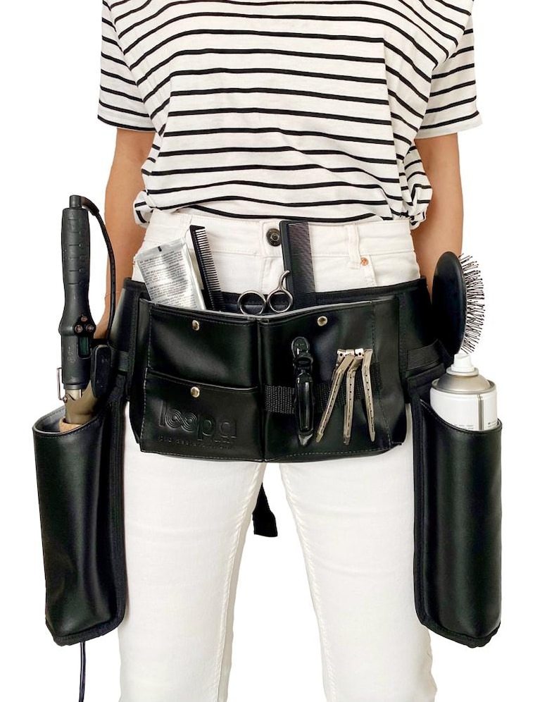 May include: Black leather stylist belt with multiple pockets and holders for tools, including a curling iron, shears, combs, and hairspray. The belt is worn by a person with white pants and a striped shirt.