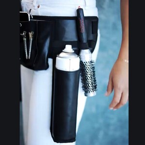 May include: A black leather hairstylist belt with a white hairspray can, a black and red hair styling tool, and a silver round brush.