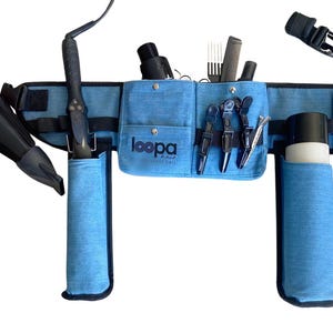 May include: A blue canvas stylist belt with multiple pockets and loops for holding hair styling tools. The belt has a black adjustable strap and is equipped with holders for a hair dryer, curling iron, and hair brushes.