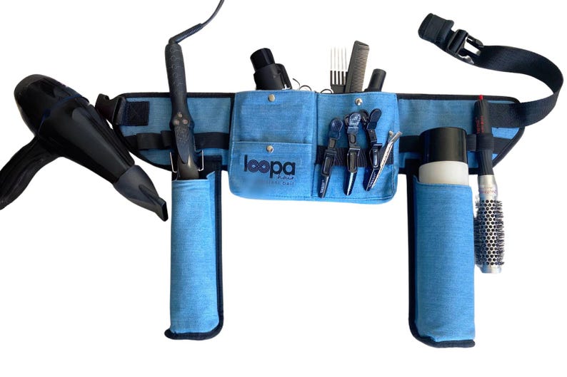 May include: A blue canvas stylist belt with multiple pockets and loops for holding hair styling tools. The belt has a black adjustable strap and is equipped with holders for a hair dryer, curling iron, and hair brushes.