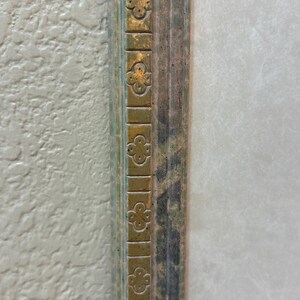 May include: Close-up of a section of a decorative picture frame. The frame has a gold-colored trim with a repeating clover-like design. The inner frame has a distressed, multi-colored appearance with shades of green, brown, and blue.