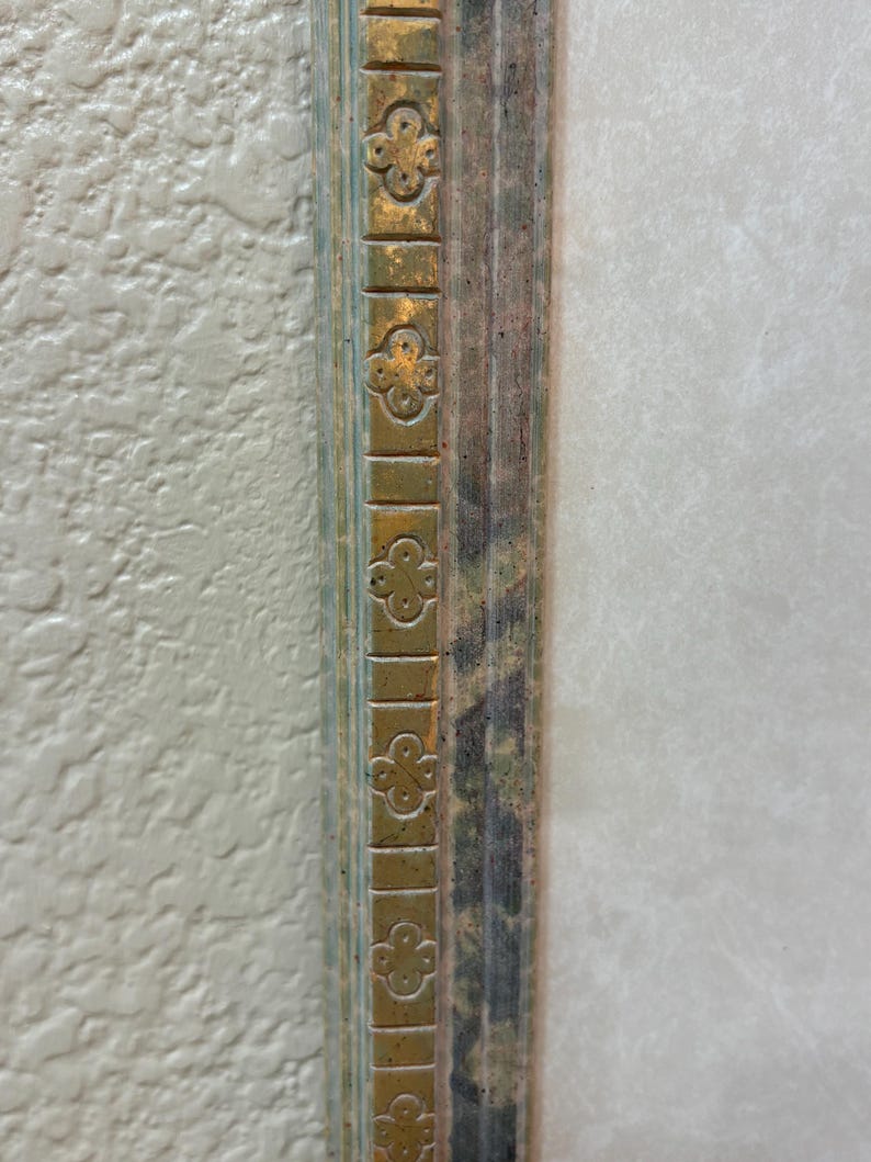 May include: Close-up of a section of a decorative picture frame. The frame has a gold-colored trim with a repeating clover-like design. The inner frame has a distressed, multi-colored appearance with shades of green, brown, and blue.