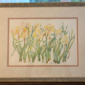 May include: Watercolor painting of yellow daffodils with green stems and leaves. The artwork is framed in a gold-toned frame with a cream-colored mat. The painting is signed in the lower right corner.