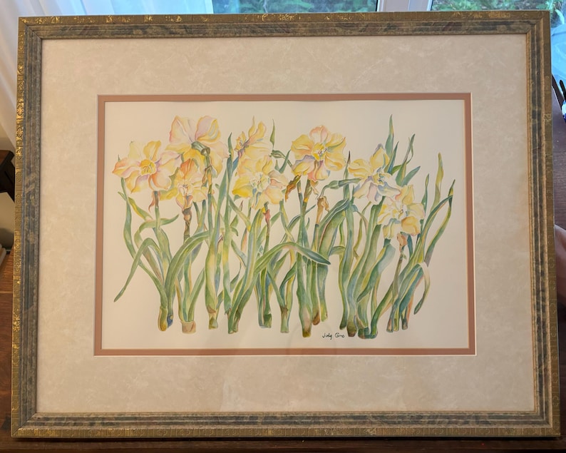May include: Watercolor painting of yellow daffodils with green stems and leaves. The artwork is framed in a gold-toned frame with a cream-colored mat. The painting is signed in the lower right corner.