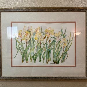 May include: Watercolor painting of daffodils in a gold-toned frame. The artwork features yellow and white flowers with green stems and leaves. The painting is set against a white background with a pink border.