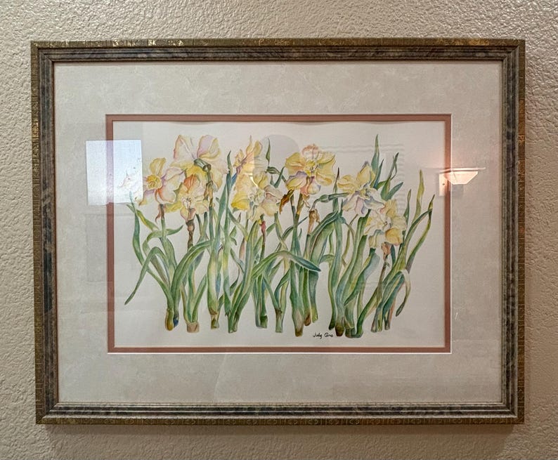 May include: Watercolor painting of daffodils in a gold-toned frame. The artwork features yellow and white flowers with green stems and leaves. The painting is set against a white background with a pink border.