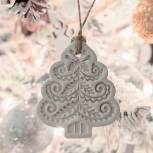 Puede incluir: Adorno de árbol de Navidad de cerámica blanca con un diseño detallado. El adorno está colgado de una cuerda de yute marrón en un árbol de Navidad blanco.